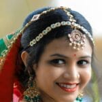Smiling Indian bride in colorful traditional attire with henna and jewelry, captured outdoors.