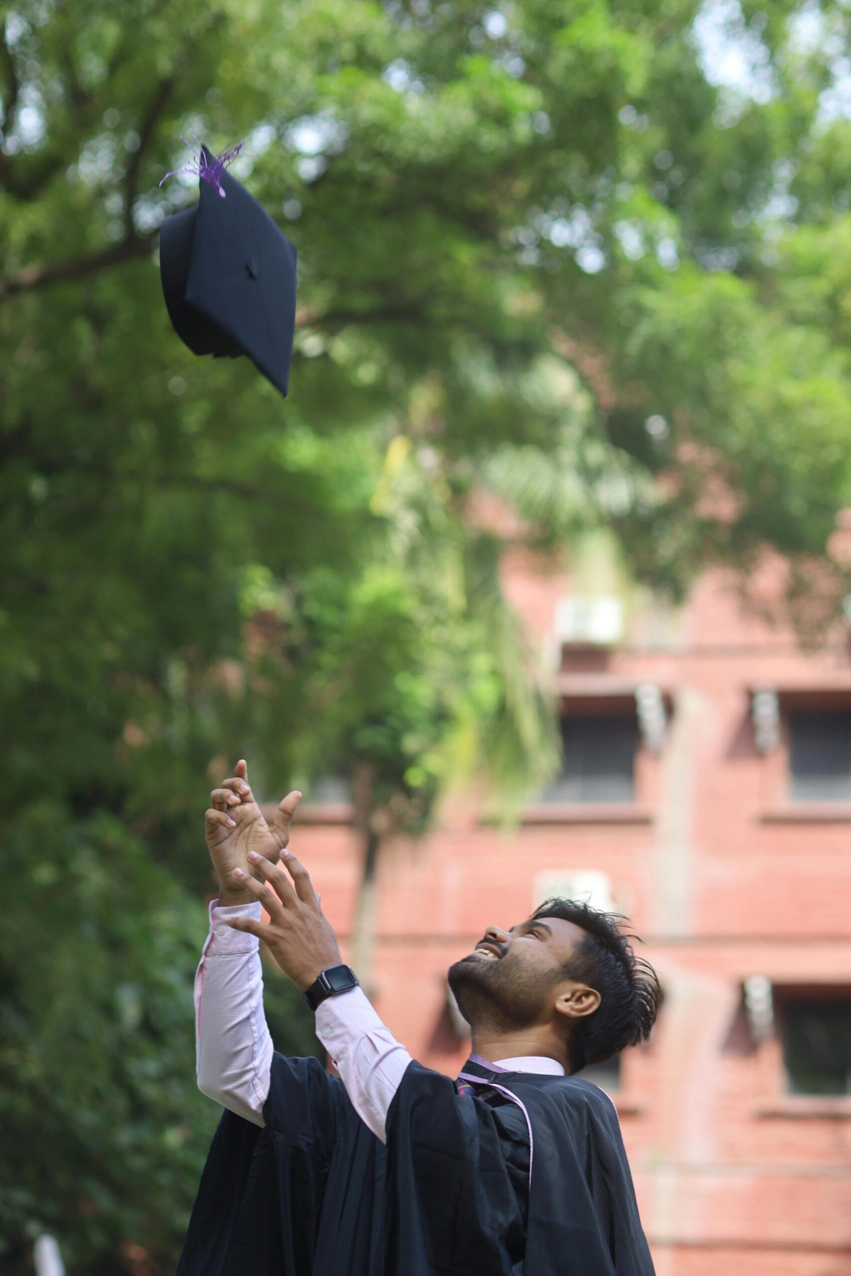 A happy graduate throws his mortarboard skyward in celebration outdoors on a sunny day.